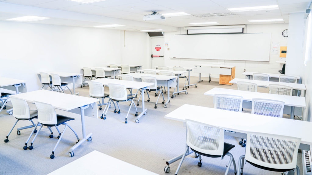 Salle épurée aux murs blancs avec un tableau blanc, des tables de travail pour deux personnes et des chaises.
